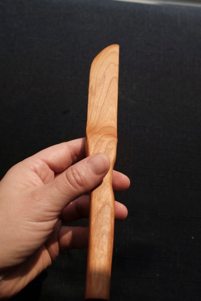 Hand holding a brown wooden knife showing the blade on its side against a black background.