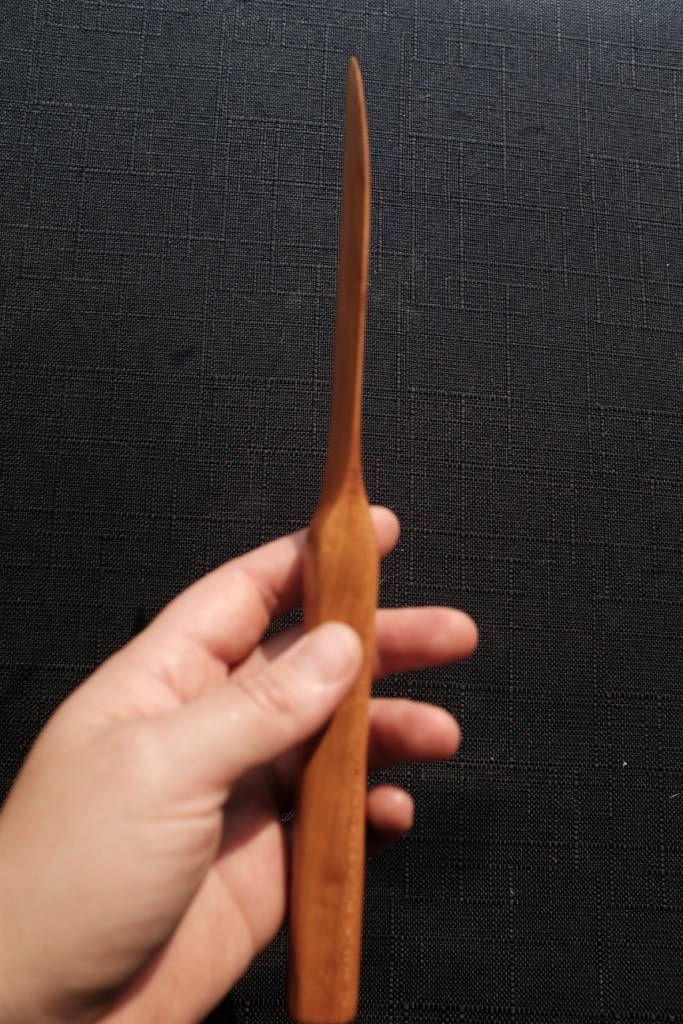 Hand holding a brown wooden knife showing the blade against a black background with the background more in focus than the knife.