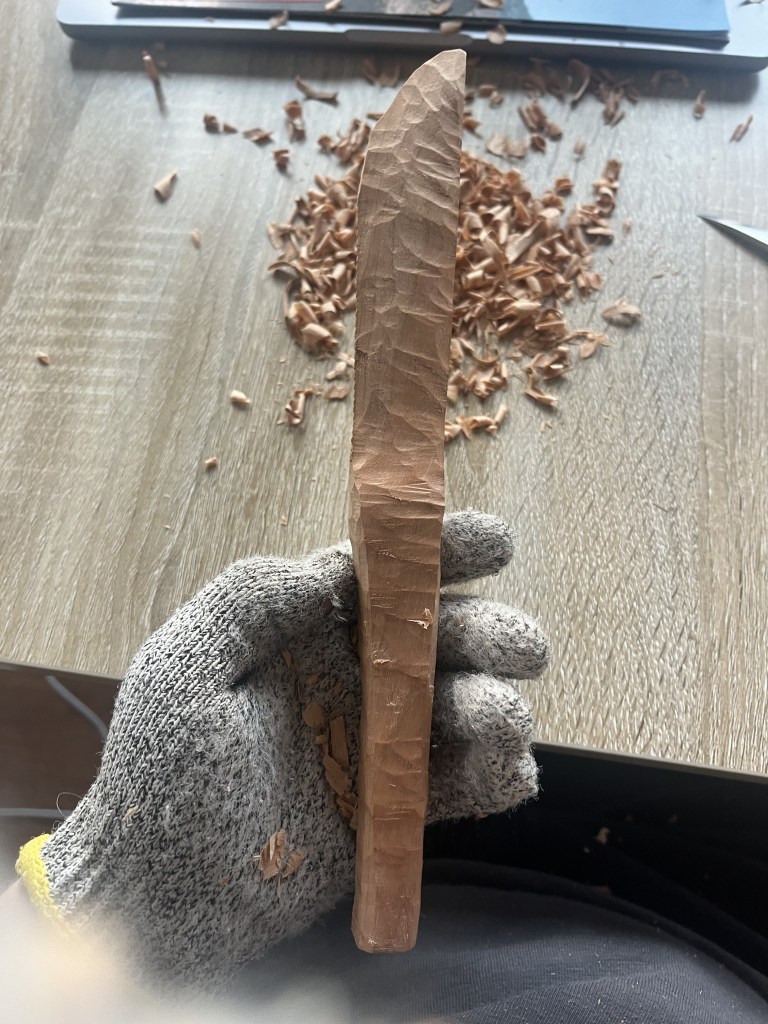 Hand with a glove on holding a wooden knife mid carving with the blade on its side.