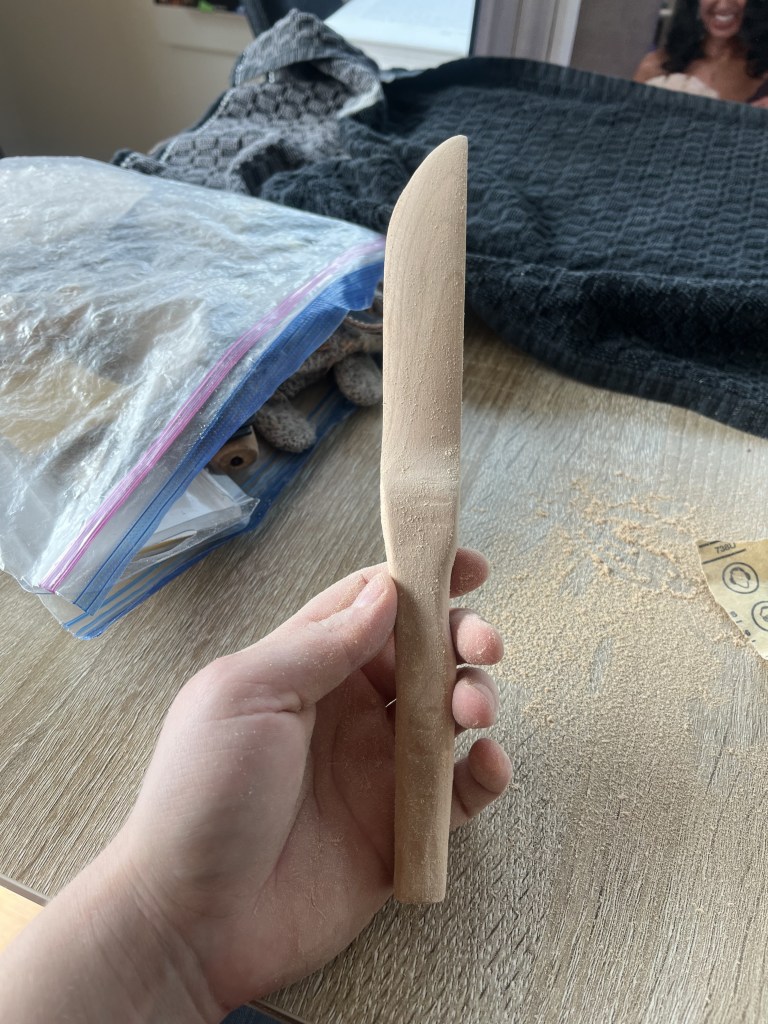 Hand holding a wooden knife showing the blade on its side in the middle of a sanding session.