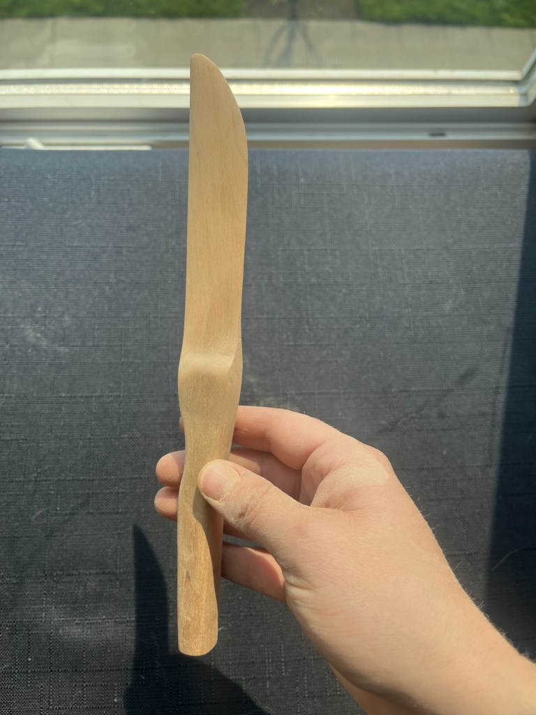 Hand holding a more white wooden knife before it's been sealed showing the blade on its side against a black background.