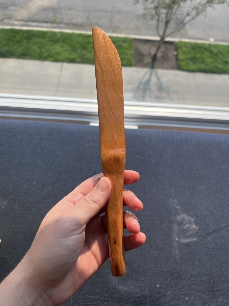 Hand holding a brown wooden knife showing the blade on its side against a black background.