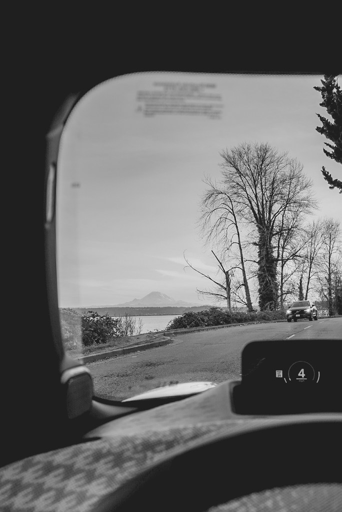 Black and white view of Mount Rainier across a lake in the distance shown from inside a car as another car approaches.